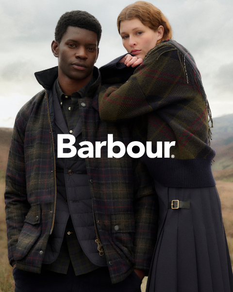 Un homme et une femme en vestes Barbour à carreaux posent en extérieur, proches l'un de l'autre, devant un ciel nuageux et un paysage vallonné. Le logo Barbour est placé au centre.