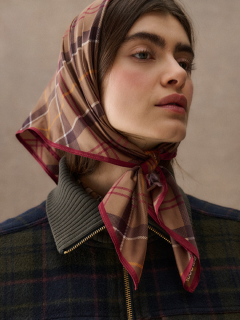 Une femme avec un foulard à carreaux noué sous son menton, porte une veste à carreaux foncée zippée et regarde légèrement vers le haut devant un fond neutre.