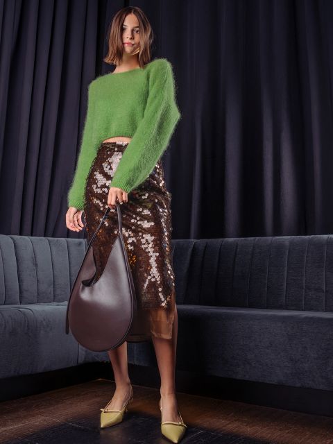 Une femme pose dans un pull vert moelleux, une jupe midi marron à paillettes et des escarpins jaune clair, et tient un grand sac hobo en cuir marron foncé devant un rideau sombre.