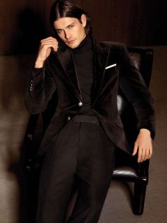 Un homme aux longs cheveux bruns est assis dans un fauteuil sombre et porte un costume en velours noir, un pull à col roulé noir et une pochette blanche.