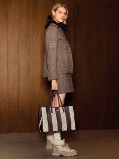 Une femme dans une tenue à carreaux tient un sac fourre-tout à carreaux à la main, se tient à l'intérieur devant un mur en bois et porte des bottes à lacets.