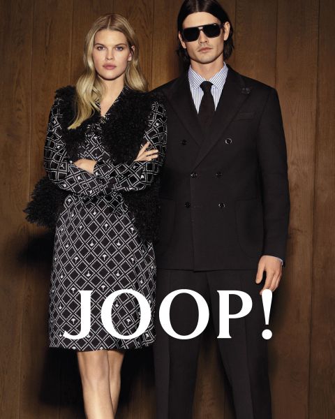 Une femme dans une robe à motifs et un homme dans un costume noir se tiennent côte à côte devant un mur en bois, sur lequel est écrit « JOOP ! » en grosses lettres blanches.
