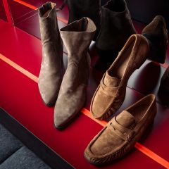 Deux paires de chaussures en daim marron - des bottines et des mocassins - exposées sur une étagère rouge brillante, dans laquelle se reflètent leurs reflets.