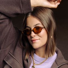Une femme aux cheveux bruns, portant des lunettes de soleil à monture écaille de tortue, une veste marron, un haut violet et des colliers dorés, et levant un bras en souriant.