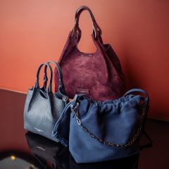 Trois sacs à main élégants - un bordeaux, un bleu marine avec une chaîne et un gris foncé - ont été présentés devant un mur rougeâtre sur une surface réfléchissante.