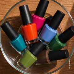 Un bol en verre, rempli de sept flacons de vernis à ongles de différentes couleurs, dont bleu, rose, orange, vert, bleu-vert, vert citron et marron.