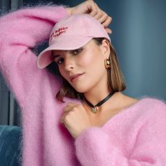 Une femme dans un pull rose moelleux et un bonnet rose pose dans une pièce ; elle porte des boucles d'oreilles dorées et un collier noir avec un pendentif cœur turquoise.