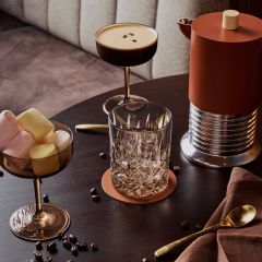 Une table avec un cocktail, un verre, un bol de marshmallows, une cafetière, une cuillère dorée, une serviette et des grains de café éparpillés.