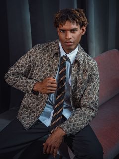 Un homme en veste à motifs, chemise et cravate rayée est assis sur un canapé rouge devant un rideau sombre, regardant la caméra avec assurance.