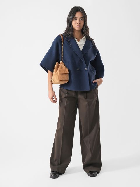 Vor einem schlichten weißen Hintergrund steht eine Frau in einem marineblauen Blazer, einer weißen Bluse, einer braunen Hose mit weitem Bein und schwarzen Schuhen und hält eine hellbraune Handtasche in der Hand.