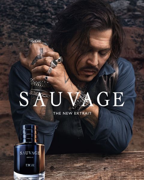 Un homme tatoué et orné de bijoux est appuyé sur une table à côté d'une bouteille de parfum « Dior Sauvage Extrait » devant un décor extérieur accidenté.