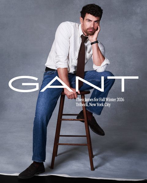 Un homme en chemise rayée, cravate et jean pose sur un tabouret en bois devant un fond gris. Texte : « GANT, Theo James pour l'automne/hiver 2026, Tribeca, New York City. »