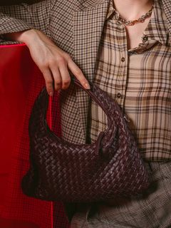 Une personne en costume à carreaux tenant un sac à main tissé marron foncé ; un fond rouge est partiellement visible en arrière-plan.