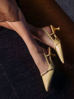 Une femme portant des escarpins jaunes à bout pointu et à lanières est assise sur un plancher en bois foncé ; sa jupe marron transparente est partiellement visible.