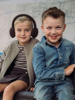 Deux petits enfants sont assis côte à côte, une fille avec des cache-oreilles et une veste et un garçon en chemise et jean en denim ; tous deux sourient à la caméra.