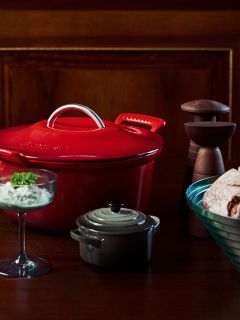 Une cocotte rouge, une petite casserole grise, un bol à dessert en verre, un bol de pain et des moulins à sel et à poivre sur une table en bois.
