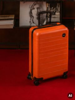 Une valise orange à poignées noires repose sur un tapis rouge devant une étagère contenant des livres et des photos en noir et blanc.