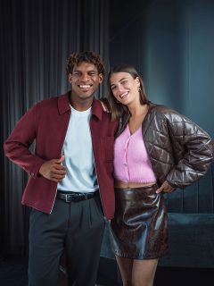 Un homme souriant dans une veste bordeaux se tient à côté d'une femme dans un haut rose et une jupe marron ; tous deux posent dans un intérieur devant un fond sombre.
