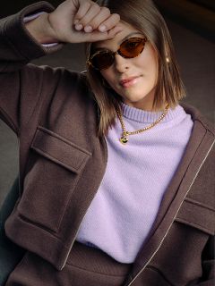 Une femme avec des lunettes de soleil marron, une veste et un pull lavande appuie son bras sur son front ; elle porte des bijoux en or et a une expression confiante.