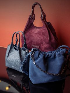 Trois sacs à main élégants - un en daim bordeaux, un en cuir bleu marine et un en daim bleu marine avec une bandoulière chaîne - sont exposés devant un fond brun-rouge.