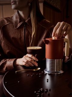 Une femme dans une veste marron est assise à une table sur laquelle se trouvent un cocktail au café, une cafetière à piston et des grains de café éparpillés.