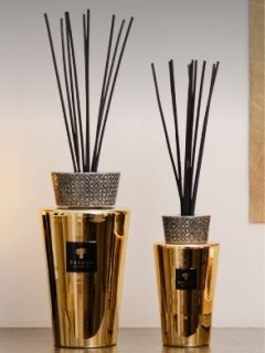 Deux vases dorés avec des parties supérieures à motifs, dans lesquels sont insérées de hautes tiges de diffuseur noires, sont posés sur une étagère devant un mur neutre.