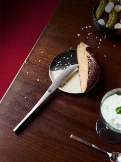 Un couteau sur une assiette avec une tranche de pain, un peu de sel et des cornichons, ainsi qu'une boisson crémeuse à côté, sur une table en bois.