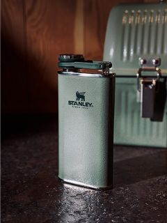 Sur un plan de travail sombre se trouve une bouteille thermos Stanley verte, et à l'arrière-plan, devant un mur en bois, on peut voir une boîte à lunch verte assortie.