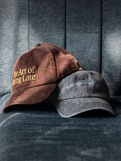 Deux casquettes sur un fauteuil en tissu bleu ; l'une marron portant l'inscription « The Art of Being Late », l'autre d'un noir délavé avec une écriture plus petite et difficilement lisible.