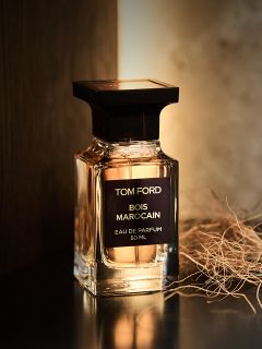 Un flacon de parfum « Bois Marocain » de Tom Ford repose sur une surface sombre à côté d'herbe séchée, tandis qu'une lumière chaude l'éclaire par l'arrière.