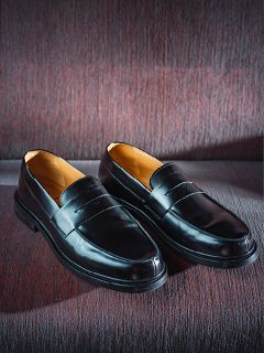 Une paire de mocassins en cuir noir brillant, posés côte à côte sur un tissu texturé de couleur bordeaux.
