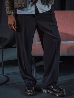 Une personne portant un pantalon noir ample, une veste à motifs et des mocassins noirs se tient à côté d'une chaise rose et d'une petite table ronde.