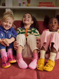Trois petits enfants sont assis sur un canapé rouge, sourient et rient ; chacun porte des vêtements colorés et des chaussures différentes et multicolores.