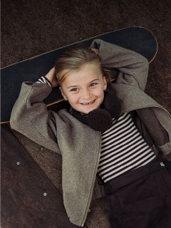 Un enfant souriant, vêtu d'une chemise rayée et d'une veste, est allongé sur un skateboard, les mains croisées derrière la tête, et regarde l'appareil photo.