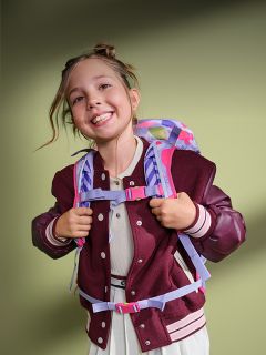 Une fille souriante avec un sac à dos et une veste bordeaux se tient devant un fond vert et a l'air joyeuse et prête pour l'école.