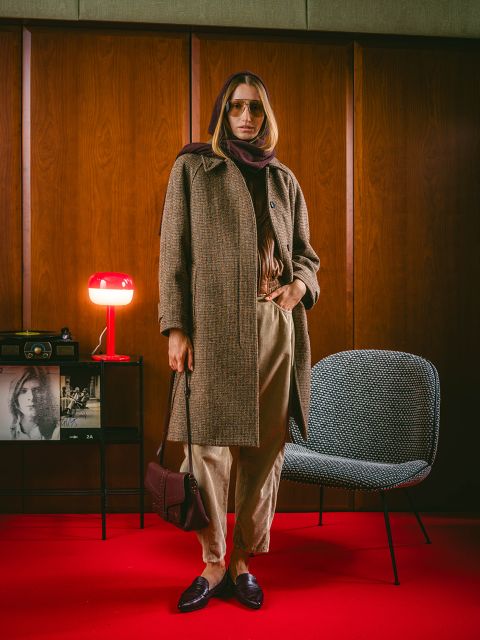 Une personne vêtue d'un manteau marron et d'une écharpe se tient dans une pièce sur un tapis rouge, avec des décorations vintage, une chaise et une lampe rouge en arrière-plan.