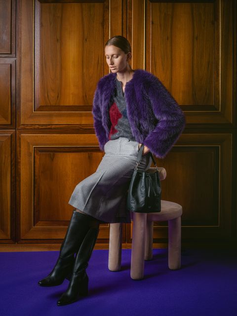 Une femme vêtue d'une veste en fourrure violette, d'une jupe grise et de bottes noires est assise avec un sac à main noir sur un tabouret devant des boiseries sur un tapis violet.