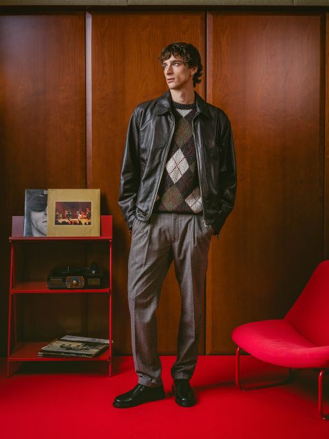 Un homme en veste de cuir et pull à carreaux se tient sur un tapis rouge à côté d'un fauteuil rouge, d'une radio rétro et d'albums devant des murs lambrissés.