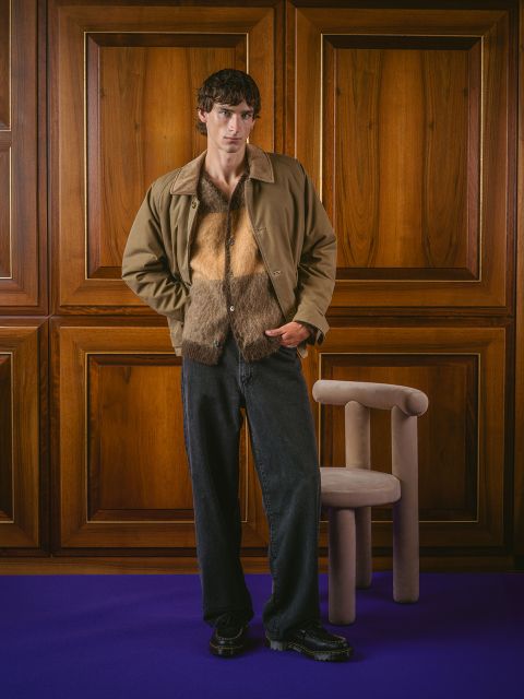 Un homme vêtu d'une veste marron, d'une chemise en fourrure et d'un jean noir se tient dans une pièce à côté d'un fauteuil beige, avec des murs lambrissés et un tapis violet.