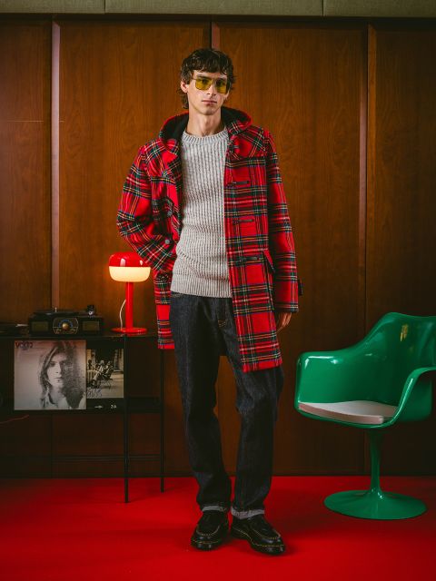 Une personne vêtue d'un manteau à carreaux rouges, d'un pull gris, d'un jean foncé et de lunettes de soleil se tient dans une pièce de style rétro avec des boiseries, une chaise verte et une lampe rouge.