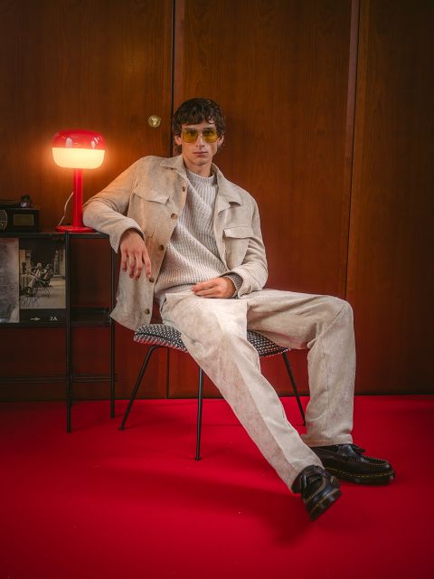 Un homme en vêtements beiges est assis sur une chaise sur un tapis rouge, derrière lui se trouvent une lampe rouge et un mur en bois. Il porte des lunettes de soleil et des chaussures noires.