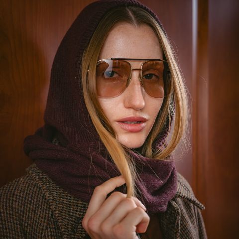 Une femme avec de grandes lunettes de soleil, une capuche-écharpe bordeaux et un manteau à carreaux marron se tient devant un fond en bois.