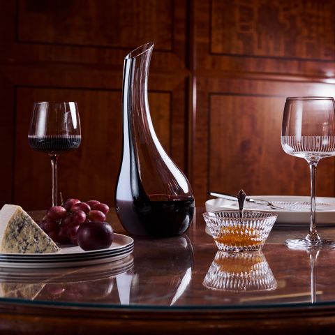 Une carafe à vin, deux verres, du fromage, des raisins et un bol de miel sur une table en bois polie, avec des assiettes visibles en arrière-plan.