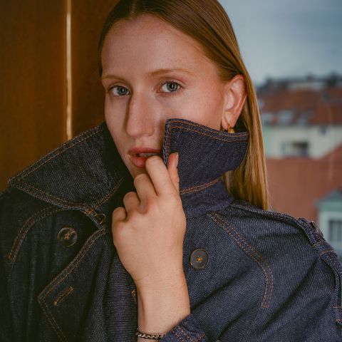 Une femme à la peau claire et aux cheveux lisses, vêtue d'une veste en jean foncé dont elle a relevé le col, se tient devant une fenêtre, avec des bâtiments visibles en arrière-plan.