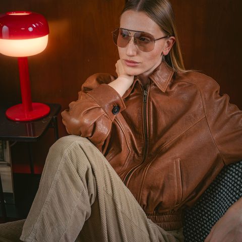 Une personne vêtue d'une veste en cuir marron et de lunettes d'aviateur est assise à côté d'une lampe rouge, l'air pensif, la main posée sur la nuque.
