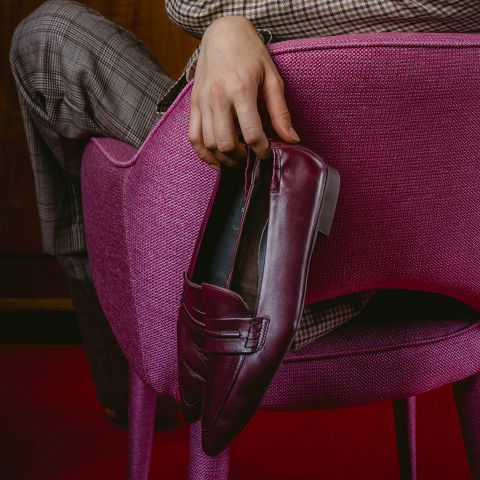 Une personne en pantalon à carreaux est assise sur une chaise rose et tient une pantoufle bordeaux sur le dossier de la chaise.