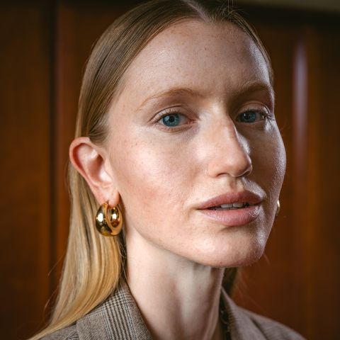 Une femme aux cheveux blonds lisses, aux créoles dorées et au maquillage discret regarde l'appareil photo, debout devant un fond en bois.
