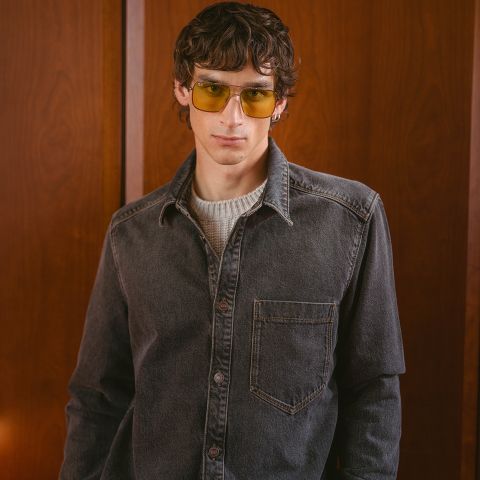 Un jeune homme aux cheveux bruns ondulés, portant des lunettes teintées de jaune, une chemise en jean grise et un pull blanc, debout devant un fond lambrissé de bois.