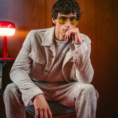 Un homme en costume de velours côtelé beige et lunettes de soleil jaunes est assis sur une chaise et pose pensivement, tandis qu'une lampe rouge est posée sur une table d'appoint à côté de lui.