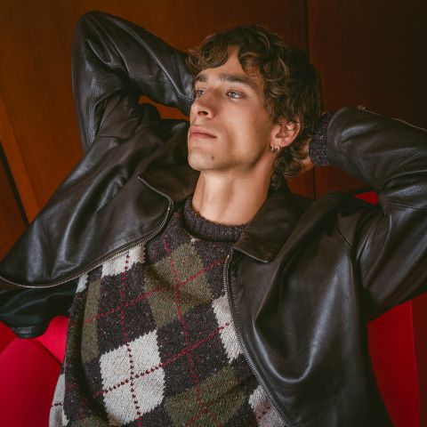 Une personne vêtue d'une veste en cuir et d'un pull Argyle est assise dans un fauteuil rouge, les mains croisées derrière la tête, et regarde pensivement vers le haut.
